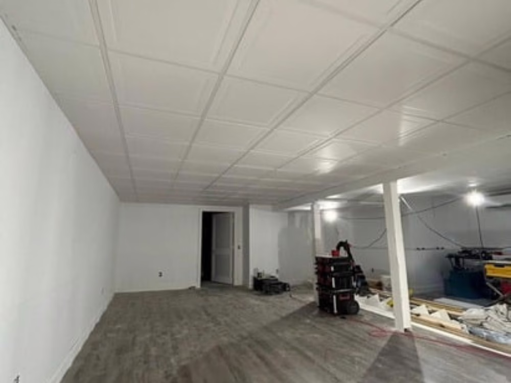 Suspended drop ceiling installation in a finished basement with white tiles and new flooring.